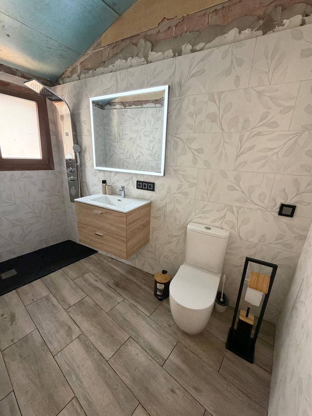 Baño reformado con plato de ducha negro, columna de hidromasaje, mueble de madera y espejo LED